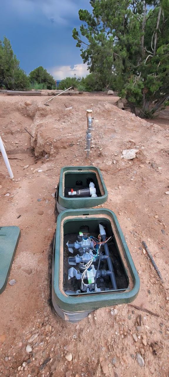 Open irrigation valve boxes with sprinkler valves, wiring, and backflow components in a dry yard